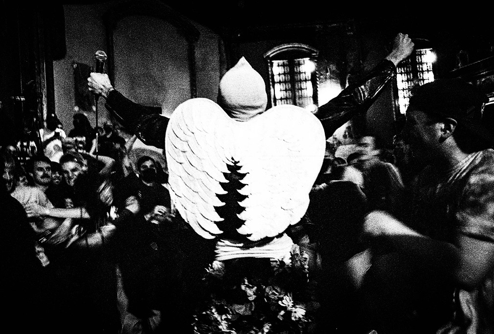 Image 1: Singer of the band Goon stands in front of the crowd at Convulse Records Fest, arms outstretched with balled fists. They are wearing a balaclava, leather jacket, costume angel wings, and ghillie-style pants covered in fake flowers. Photo credit: Copyright of this image is retained by Joe Lacey (@joexlacey) and used by the authors with permission.